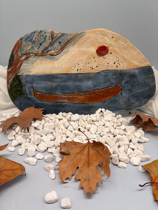 This handcrafted ceramic art plate captures a serene lakeside scene, featuring a lone boat resting on calm blue waters beneath a warm red sun. The textured surface and painterly glazes in earthy browns, blues, and creams evoke a sense of quiet reflection and natural harmony.  Perfect for wall display or as a tabletop accent, this piece embodies peaceful stillness and the timeless 