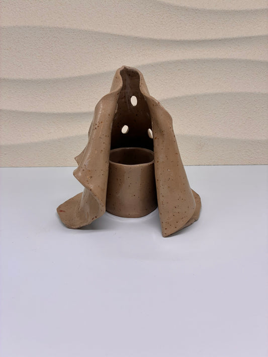 This handcrafted ceramic candle holder features an organic, folded form with carefully placed openings that allow the candlelight to softly glow through. Its sculptural silhouette evokes the feeling of draped clay or natural rock formations, creating a warm and atmospheric presence when lit. Designed to hold a candle securely inside, it functions as both a light source and a decorative art object. Each piece is handmade, with subtle variations that highlight its unique, artisanal character.