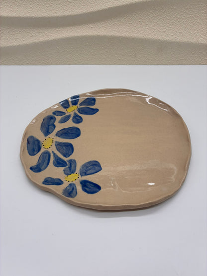 This handcrafted ceramic round plate is adorned with small hand-painted blue flowers, creating a delicate and timeless pattern across the surface. Each plate is individually painted by hand, making every piece unique. Designed exclusively for serving, it combines artisanal craftsmanship with a refined decorative touch, adding elegance and character to the table.