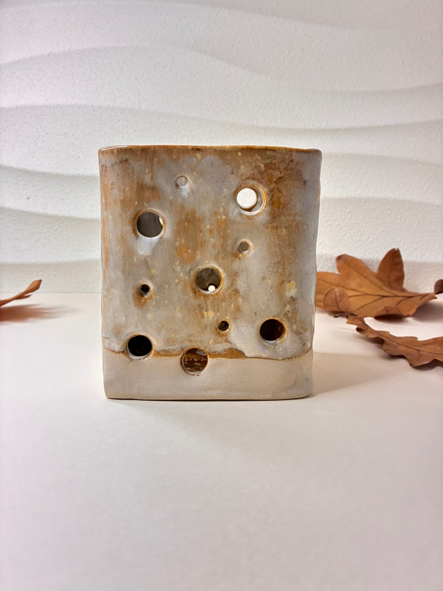 This handcrafted ceramic candle holder features a short, rounded form with decorative perforations, designed to hold a candle and create a warm, ambient glow. When lit, the light shines through the openings, casting beautiful patterns of shadows and light. Each piece is carefully made by hand, with subtle variations that highlight its artisanal character. Perfect for cozy evenings, table settings, or as a decorative accent, it combines functionality with sculptural charm.