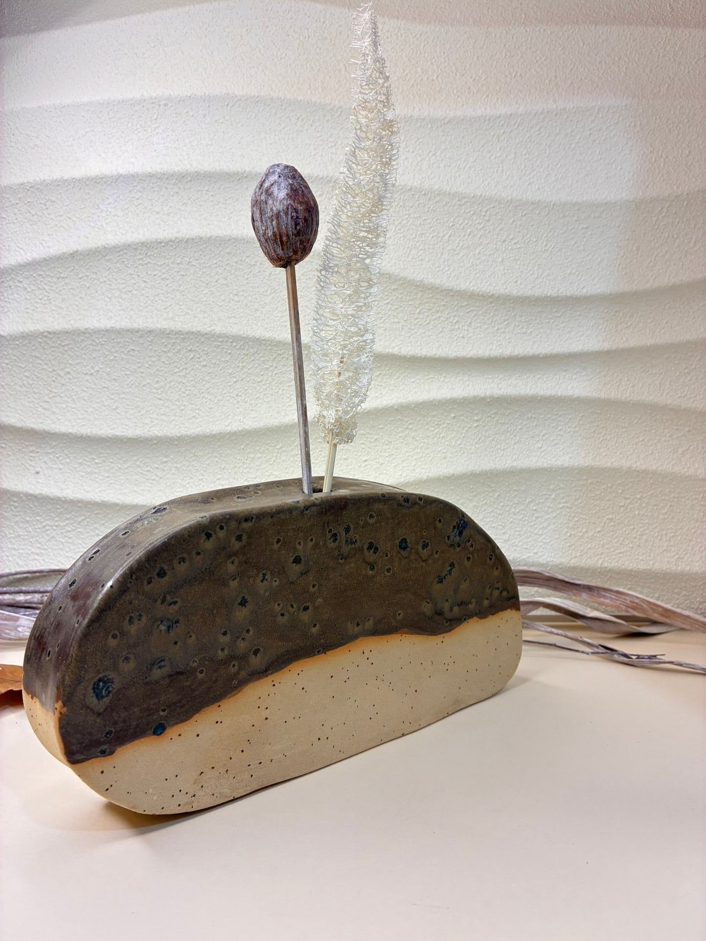 Handcrafted as a sculptural ceramic vase, this piece features a deep, earthy palette with a rich, iron-toned glaze marked by natural pitting and subtle surface variation. Its rounded, hollowed form evokes volcanic stone and ancient vessels shaped by time and heat. The matte-to-satin finish and organic markings highlight the firing process, ensuring each piece is truly one of a kind. Fully functional, it can hold water for fresh flowers, while also serving as a vase for dried stems or standing alone as a gro