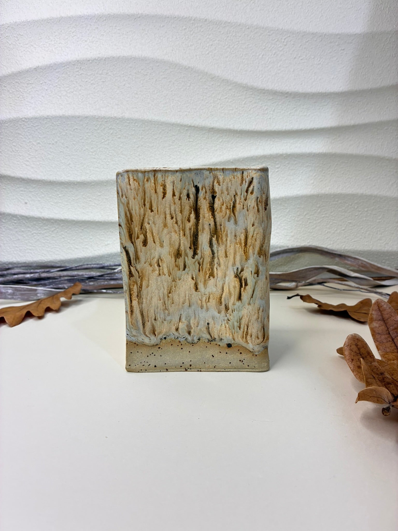 Handmade with care, this ceramic vessel features a softly squared form and a richly textured glaze reminiscent of weathered driftwood and cascading mineral stains. Vertical flows of warm cream, sand, and charcoal tones create an organic, one-of-a-kind surface, while the speckled stone base grounds the piece with a natural, earthy feel.