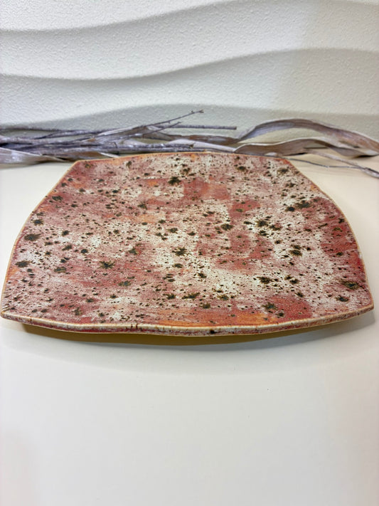 The Terra Bloom Plate is a celebration of natural texture and earthy warmth. Its soft, square form is glazed in a muted rose tone, speckled with rich brown and black markings that resemble organic moss patterns on sunbaked clay. The slightly curved edges and handcrafted finish give it a grounded yet elegant feel. Ideal for serving or display, this piece evokes the beauty of aged stone and wild landscapes — a perfect fusion of rustic artistry and refined simplicity.