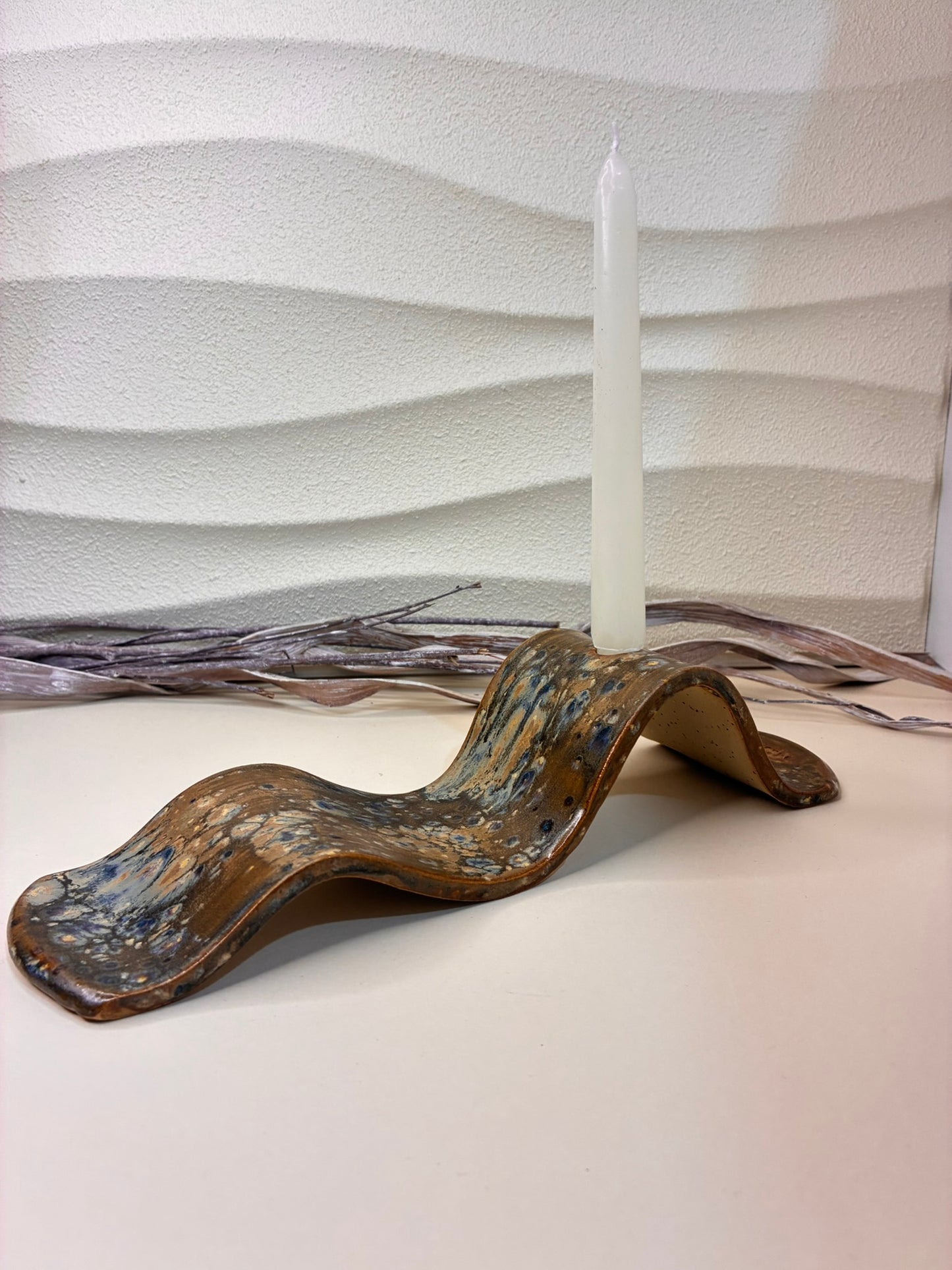 This handcrafted elongated ceramic form features a soft, wavy silhouette with a single opening, designed to hold a candle or a dried stem. Its flowing, curved shape gives it a sculptural presence, making it both a functional object and a decorative statement. Each piece is individually made by hand, with subtle variations that highlight its artisanal character. Whether used with a flame or as a minimal vessel for dried botanicals, it brings a sense of movement, calm, and organic elegance to any space.