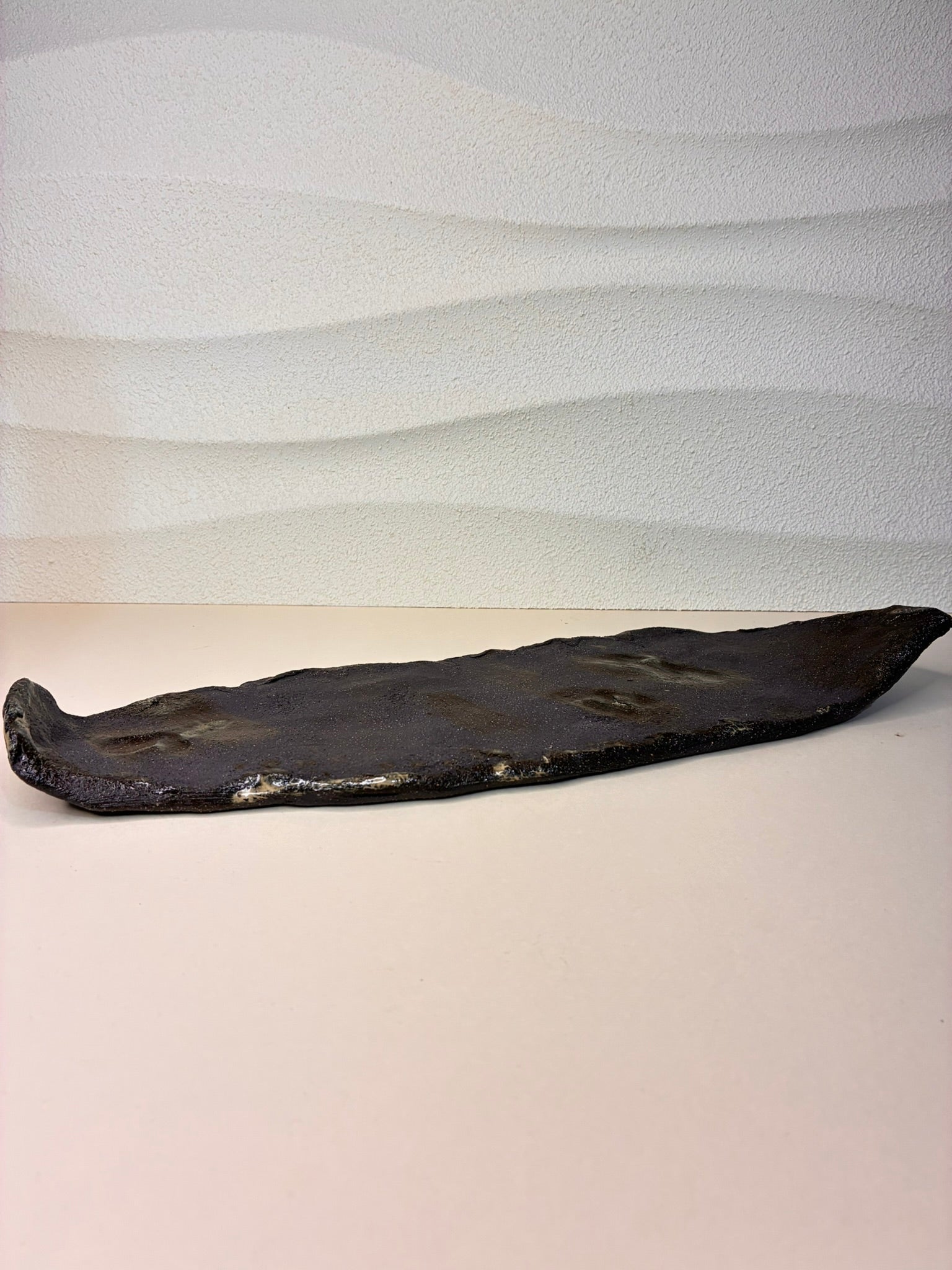 This handcrafted elongated tray is made from black stoneware and features gently raised edges with a volcanic stone-inspired texture. Its organic form and raw, earthy surface make it a striking decorative piece, perfect for displaying objects, holding trinkets, or adding sculptural elegance to any space. Each tray is unique, with subtle variations that highlight its artisanal character. A fusion of natural texture and minimalist design, it brings a sense of quiet artistry and grounded sophistication to your