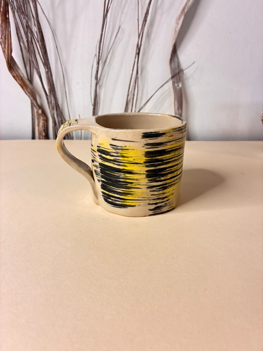 A handmade ceramic mug crafted from natural white clay and decorated with expressive black brushstrokes layered with warm yellow accents. Each mark is applied by hand, giving the surface a dynamic, painterly texture and making every mug unique. The organic shape and raw finish highlight the beauty of imperfection, creating a functional piece of art for everyday coffee or tea moments.