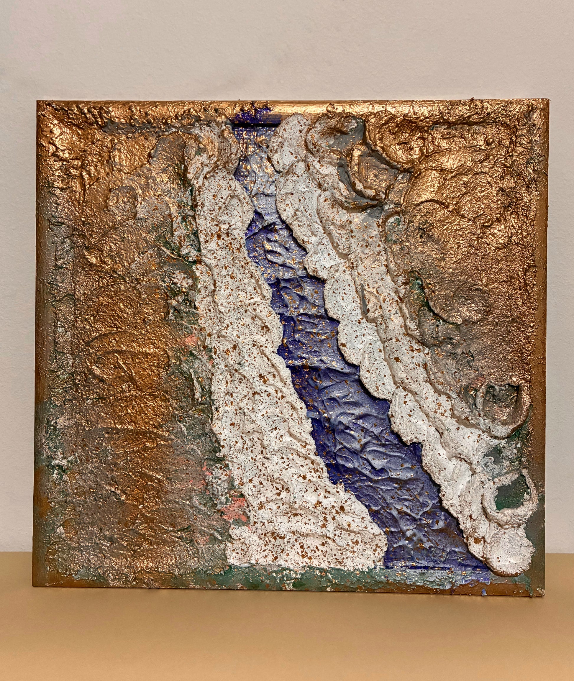 Mixed-media artwork crafted with textured materials like plaster and clay. It evokes a river weaving through a desert landscape, with layered textures suggesting movement, depth, and the meeting of natural elements.