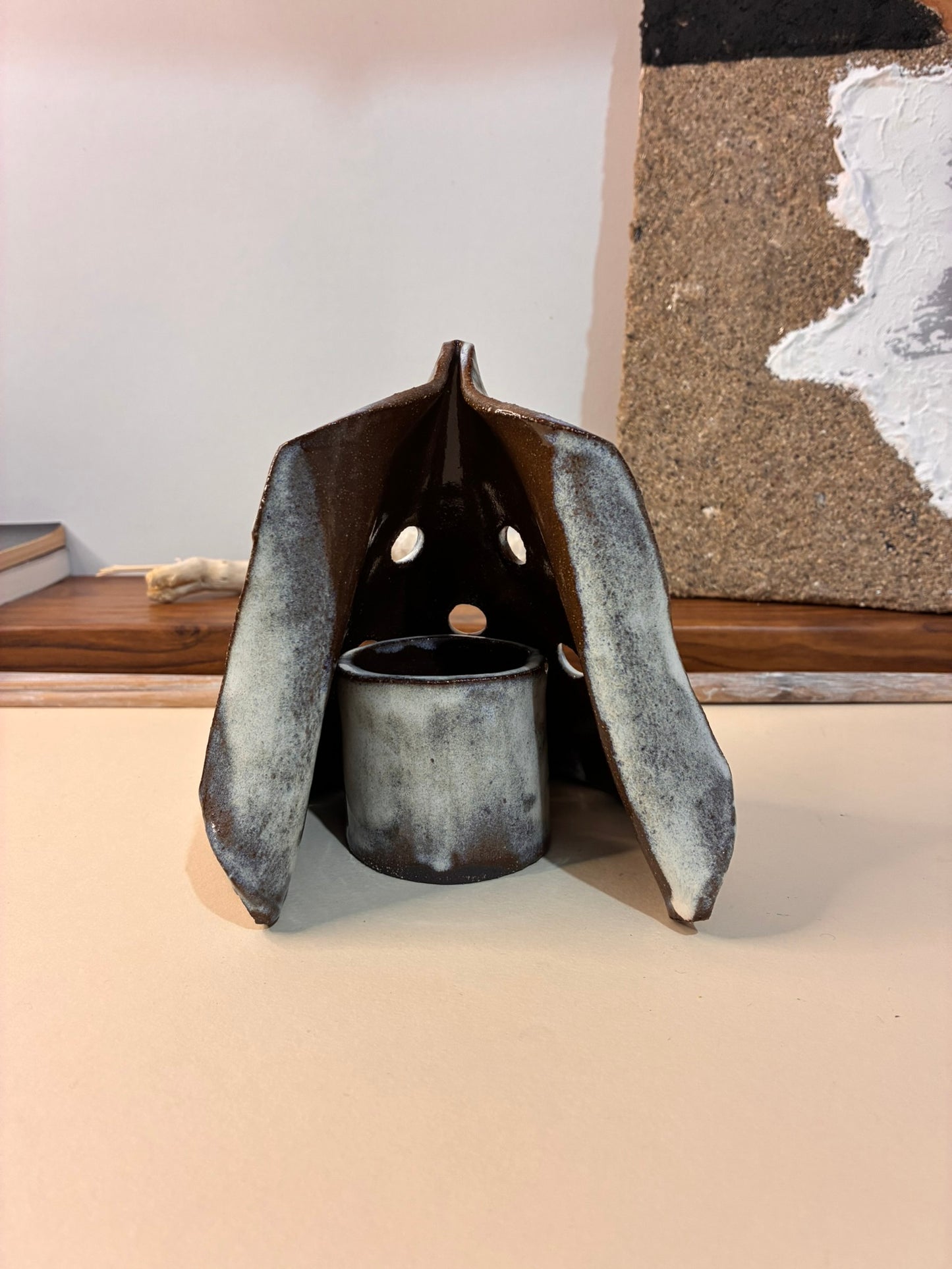 Bring warmth and quiet character into your space with this handcrafted ceramic candle holder. Shaped like a small shelter, its folded form softly cradles a tealight, allowing the flame to glow gently through the cutout openings. The natural earth-toned glaze and organic texture make each piece one of a kind.

Perfect for cozy evenings, meditation corners, or minimalist interiors, this candle holder creates a calming ambiance while standing beautifully on its own as a sculptural object.